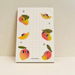 Puede incluir: Un bloc de notas con un patrón de cuadrícula e ilustraciones de mangos de dibujos animados con caras y hojas verdes. Los mangos son amarillos y rojos, algunos con sombreros rosas. El bloc de notas tiene la palabra "bloemkolie" impresa y pequeñas estrellas.