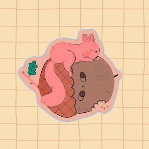 Op de afbeelding: Een gestanste sticker met een roze eekhoorn die rust op een grote bruine eikel. De eikel heeft een getextureerd oppervlak met een rasterpatroon aan één kant en een klein groen blaadje aan een dun steeltje aan de linkerkant.