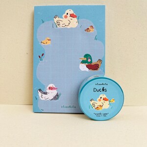 May include: A blue notepad and washi tape set featuring duck illustrations. The notepad has a grid pattern and duck drawings. The washi tape roll is labeled "Ducks" and "washi tape" with measurements of 1.5cm x 10m.