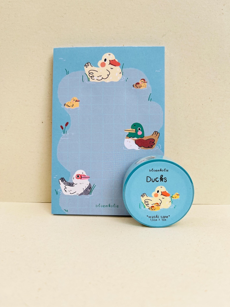 May include: A blue notepad and washi tape set featuring duck illustrations. The notepad has a grid pattern and duck drawings. The washi tape roll is labeled "Ducks" and "washi tape" with measurements of 1.5cm x 10m.