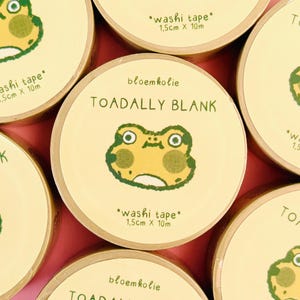 Op de afbeelding: Meerdere rollen washi tape met een lichtgeel etiket met een groene kikkerillustratie. De tekst luidt bloemkolie, TOADALLY BLANK en washi tape 1,5cm x 10m. De kikker heeft grote ogen en ronde wangen, gecentreerd op elke ronde tape-rol.