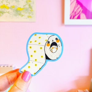 May include: A blue and white sticker of a cartoon animal peeking out of a roll of toilet paper. The animal has a white body, black eyes, and orange cheeks. The toilet paper is white with yellow dots.