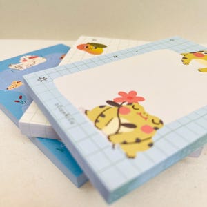 May include: A stack of square notepads with cartoon illustrations. The top notepad has a light blue grid pattern and a yellow cartoon character with a red flower. Other notepads have similar designs with different characters and colors.