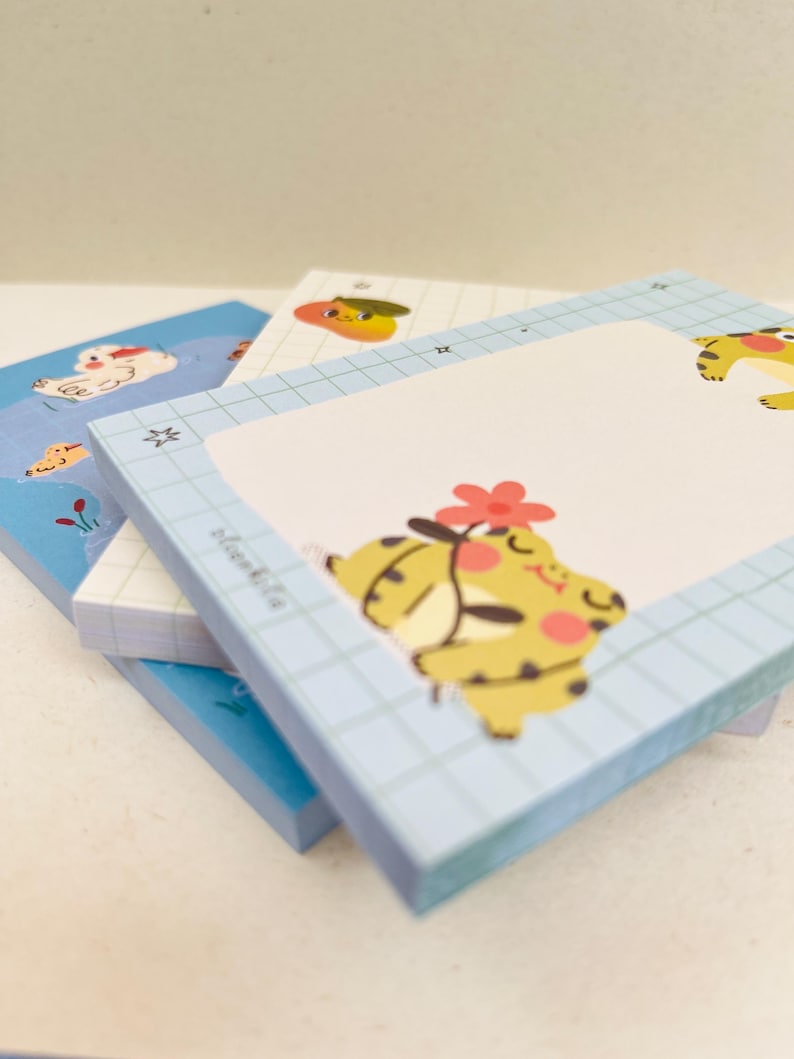 May include: A stack of square notepads with cartoon illustrations. The top notepad has a light blue grid pattern and a yellow cartoon character with a red flower. Other notepads have similar designs with different characters and colors.