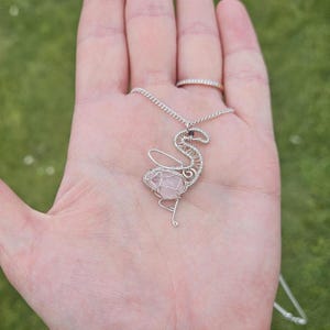 Pode incluir: Um pingente de fio de prata em forma de flamingo com uma pedra de quartzo rosa no centro. O pingente está pendurado em uma corrente de prata.