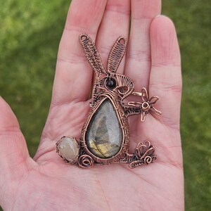 May include: A copper wire wrapped pendant featuring a labradorite gemstone in the shape of a rabbit. The rabbit is holding a small flower in its paw and has a smaller gemstone on its tail.