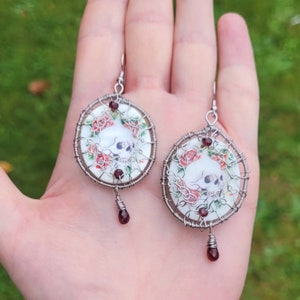 May include: A pair of silver wire wrapped earrings with a white porcelain skull and floral design. The earrings have a dangling red gemstone.