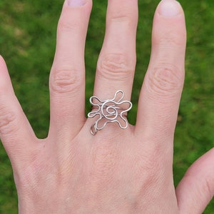 May include: A silver wire ring with a swirl design. The ring is on a person's finger.