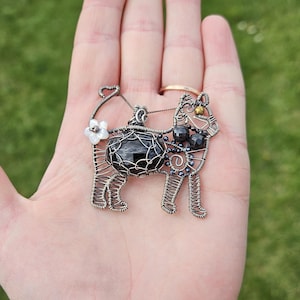 May include: A silver wire-wrapped pendant featuring a black gemstone, black beads, and a white flower accent. The pendant is shaped like a cat.
