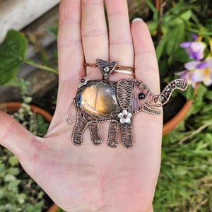 May include: A handcrafted copper wire elephant pendant with a labradorite stone center. The pendant features a detailed wire-wrapped design, small flower accents, and a dark gemstone eye. The elephant is held in a hand.