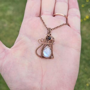 May include: A copper wire-wrapped cat pendant necklace featuring a pearlescent, iridescent stone. The cat pendant has a detailed wire frame with a small black bead accent. The necklace is displayed on a hand.