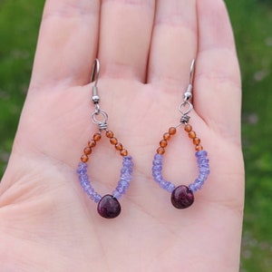May include: A pair of dangle earrings with a teardrop shape. The earrings are made with small, round beads in shades of amber and purple. The earrings are finished with a larger, dark purple gemstone.