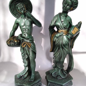May include: A pair of green ceramic figurines depicting a man and a woman in traditional Asian clothing. The man is wearing a straw hat and holding a basket. The woman is holding a lantern. Both figurines are standing on a green base.