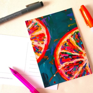 Könnte beinhalten: Eine Postkarte mit einem lebendigen Gemälde von zwei Orangenscheiben mit roten Umrissen und bunten Segmenten auf türkisblauem Hintergrund. Ein schwarzer Stift, ein rosa Stift und ein orangefarbener Marker sind ebenfalls abgebildet.
