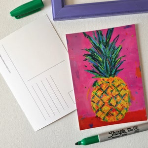 May include: A vibrant postcard featuring a colorful painting of a pineapple against a pink background. The pineapple is rendered in shades of yellow, orange, green, and blue. A blank postcard and a green marker are also visible.