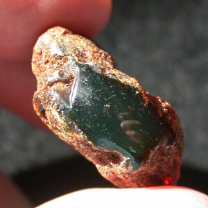 May include: A dark green gemstone with a rough, brown exterior. The gemstone is irregularly shaped and has a smooth, polished surface.