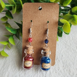 May include: A pair of handmade earrings featuring miniature glass potion bottles. One bottle contains red liquid and is labeled "Health", the other contains blue liquid and is labeled "Mana". Each bottle is topped with a cork and hangs from a silver hook.