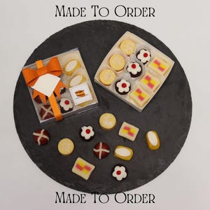 May include: Two square boxes filled with assorted marzipan confections, including flower-shaped, rectangular, and round pieces. One box features an orange ribbon and a tag reading Marzipan Artisan. Additional loose marzipan treats are scattered nearby.