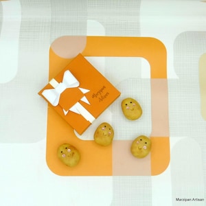 May include: A small orange gift box with a white bow and the text "Marzipan Artisan" on the top. Four small marzipan potatoes with painted faces are arranged around the box on a white and orange background.