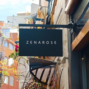 May include: A black rectangular shop sign with the name "ZENAROSE" in white lettering. The sign is suspended from a black metal bracket. The building has a textured, light-coloured facade, with colourful plants and flowers.