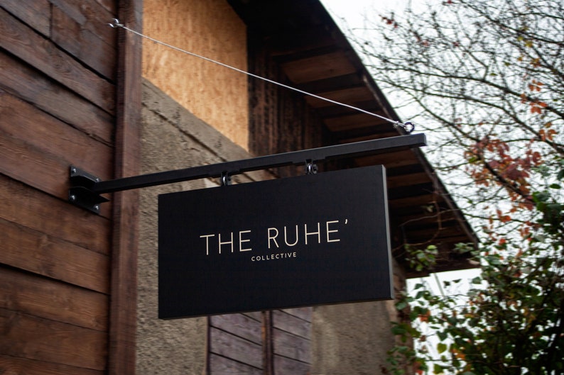 May include: A black rectangular sign displaying the text "THE RUHE'" and "COLLECTIVE" in gold lettering. The sign is mounted on a black metal bracket, attached to a wooden building.