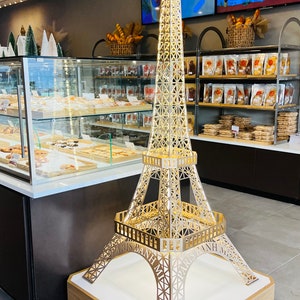 May include: A gold Eiffel Tower replica with the words "Paris Banh Mi" on the base, standing on a wooden platform in a bakery.