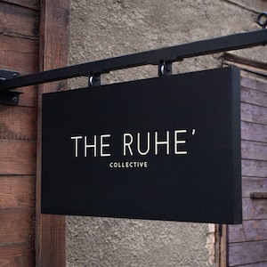 May include: A black rectangular sign with the text "THE RUHE' COLLECTIVE" in white lettering. The sign is suspended from a black metal bracket attached to a wooden wall.