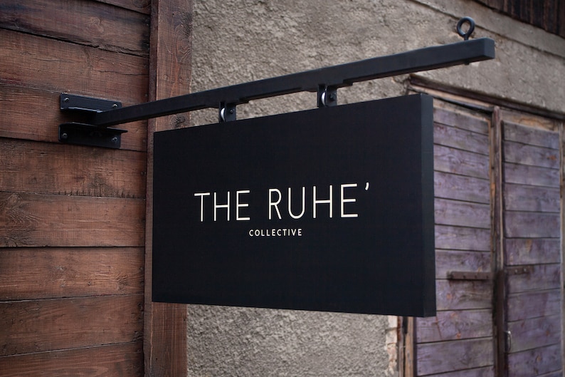 May include: A black rectangular sign with the text "THE RUHE' COLLECTIVE" in white lettering. The sign is suspended from a black metal bracket attached to a wooden wall.