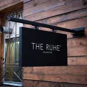 May include: A black rectangular sign with the text "THE RUHE' COLLECTIVE" in white lettering, mounted on a black metal bracket. The sign is attached to a wooden wall with horizontal planks.