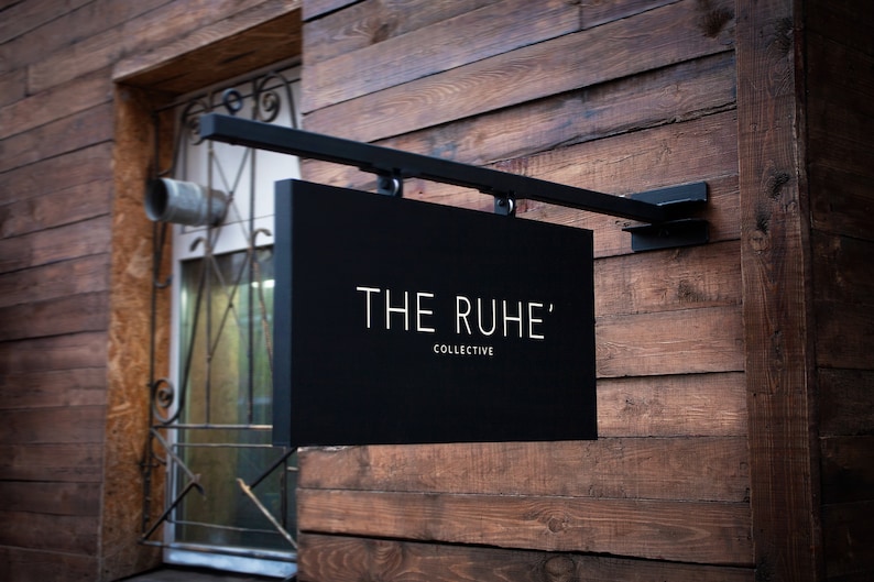 May include: A black rectangular sign with the text "THE RUHE' COLLECTIVE" in white lettering, mounted on a black metal bracket. The sign is attached to a wooden wall with horizontal planks.