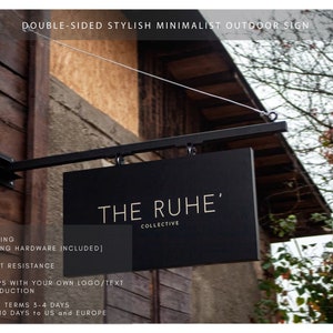 May include: A black, double-sided outdoor sign with the text "THE RUHE' COLLECTIVE" in gold lettering. The sign is suspended from a black metal bracket and cable. The sign is mounted on a wooden building.
