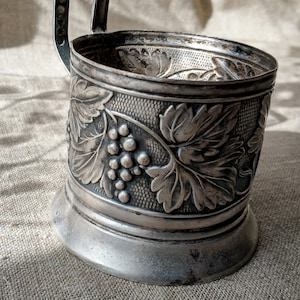 Könnte beinhalten: Silberbeschichteter Becherhalter mit einem verzierten Design aus Weinreben und Trauben. Der Halter hat einen Griff und einen abgerundeten Boden.