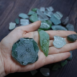 Op de afbeelding: Een hand die verschillende stukken zeeglas vasthoudt, waaronder een groot, groen stuk met een getextureerd oppervlak en een kleiner, lichtblauw stuk met een swirl-ontwerp.