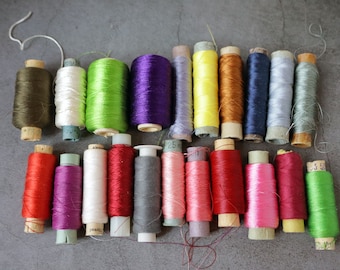 Può includere: Una collezione di bobine di filo di vari colori, tra cui verde, viola, rosso, rosa, giallo, arancione, blu e grigio. Le bobine sono disposte in due file su una superficie grigia.