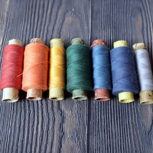 May include: Eight spools of thread in various colors, including red, orange, yellow, green, blue, and gray, are arranged in a row on a wooden surface.