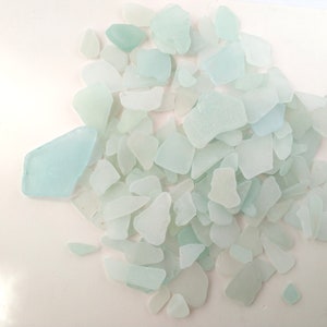Op de afbeelding: Een verzameling stukken zeeglas in diverse vormen en maten. Het zeeglas is voornamelijk lichtblauw en wit van kleur, met een matte, gladde textuur. De stukken zijn verspreid op een witte ondergrond, wat een natuurlijke, strandachtige uitstraling creëert.