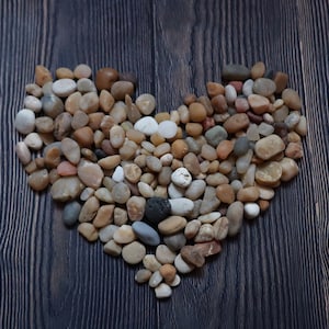 Può includere: Una forma a cuore realizzata con piccole pietre lisce, marroni, beige e grigie su uno sfondo di legno scuro.