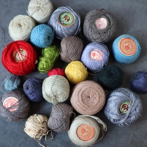 Op de afbeelding: Een verzameling kleurrijke garenbollen, sommige met labels, gerangschikt in een cirkel op een grijs oppervlak.