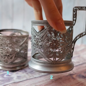 Peut inclure: Deux supports à verre à thé en métal argenté avec des motifs filigranés complexes. Le support de gauche est de forme cylindrique simple, tandis que le support de droite a une poignée et un design plus élaboré.