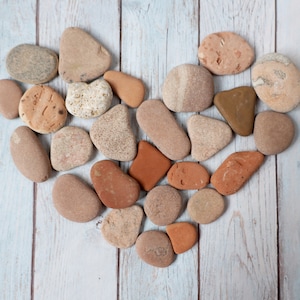 May include: A heart shape made of various smooth, rounded stones in shades of tan, brown, and reddish-orange. The stones are arranged on a light blue and white wooden surface, creating a natural and earthy aesthetic.