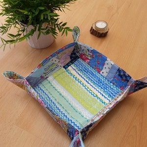 Könnte beinhalten: Ein handgefertigtes, quadratisches Stofftablett mit Patchwork-Design. Das Tablett weist eine Vielzahl von Stoffen mit blauen, grünen und floralen Mustern auf. Die Kanten sind hochgeklappt, um die Seiten zu bilden, und das Tablett steht auf einer Holzoberfläche.