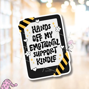 May include: Black and white sticker with yellow and black striped tape. The sticker features a Kindle e-reader with the text "Hands off my emotional support Kindle".