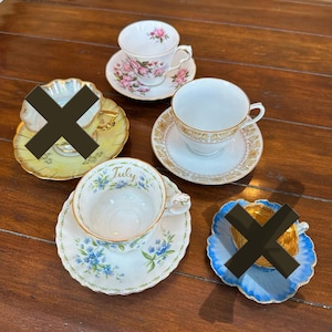 May include: Four teacups and saucers with floral and gold designs. The teacups are white with gold trim and floral patterns. The saucers are white with gold trim and floral patterns. One saucer has the word "July" written on it.