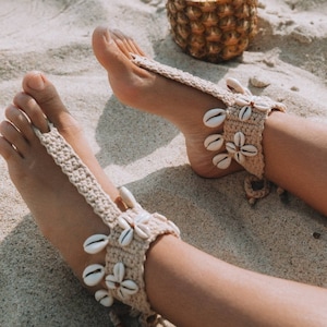 May include: Beige crochet barefoot sandals adorned with seashells. The sandals feature a toe loop and ankle strap design, with seashell accents arranged in a floral pattern. A pineapple with a straw is in the background.