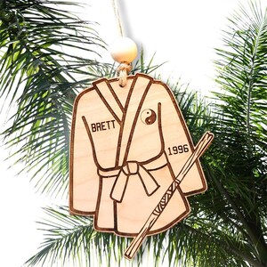 May include: A wooden ornament shaped like a karate uniform with a white belt, a stick, and the yin-yang symbol. The ornament is personalised with the name "Brett" and the year "1996".