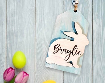 May include: A wooden Easter tag with a bunny silhouette and the name "Braylie" in script. The tag is decorated with a blue and white watercolor effect. The image also features colorful Easter eggs, tulips, and a nest.
