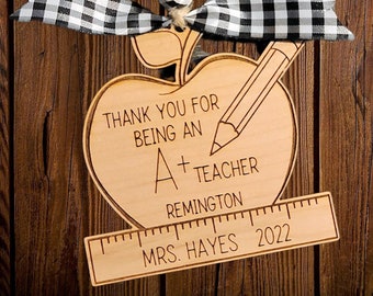 May include: Wooden apple ornament with a black and white checkered ribbon, a pencil, and the text "Thank you for being an A+ teacher Remington Mrs. Hayes 2022".