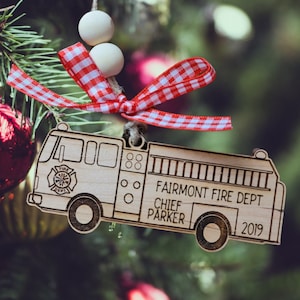 May include: Wooden Christmas ornament shaped like a fire engine. The ornament has the text "Fairmont Fire Dept, Chief Parker, 2019". It is decorated with a red and white checkered ribbon and two white wooden beads.