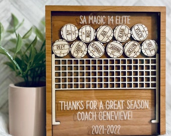 May include: A wooden shadow box with a volleyball net design and the text "SA Magic 14 Elite" and "Thanks for a great season. Coach Genevieve! 2021-2022". The box is filled with wooden volleyball-shaped tokens, each with a player's name and number.