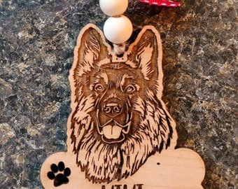 Può includere: Ornamento per cani in legno con un'incisione dettagliata del volto di un pastore tedesco. L'ornamento include un'etichetta a forma di osso con il nome "KIKI" e impronte di zampe. È adornato con un nastro rosso a pois bianchi e due perline di legno.